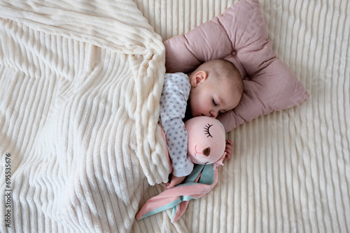 Baby with pink bunny soft toy. A girl of European appearance, wearing a light light jumpsuit, is sleeping in the crib. Sleep schedule, care and hygiene of a child up to one year old.