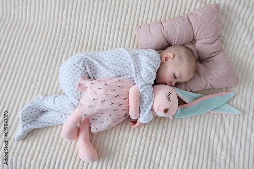 Baby with pink bunny soft toy. A girl of European appearance, wearing a light light jumpsuit, is sleeping in the crib. Sleep mode, care and hygiene.