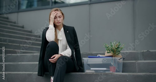 Sad woman is sitting on stairs outside an office building, wearing formal suit.