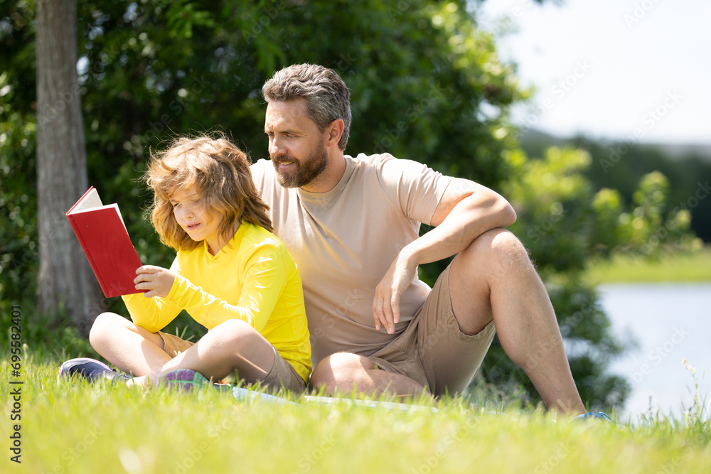Father and son enjoy spending quality time reading books outdoors. Portrait of father and son relax on green lawn in summer park and read book together. Father and his little son reading book in park. Father and son enjoy spending quality time reading books outdoors. Portrait of father and son relax on green lawn in summer park and read book together. Father and his little son reading book in park.