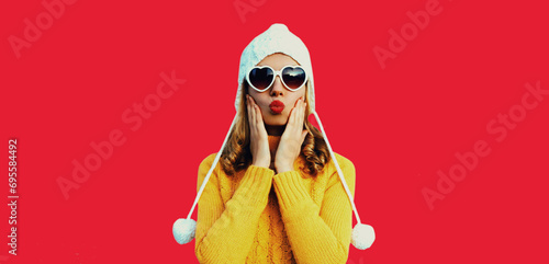 Winter portrait of beautiful woman blowing her red lips sending sweet air kiss wearing heart shaped sunglasses, yellow knitted sweater and white hat with pom pom on studio background photo