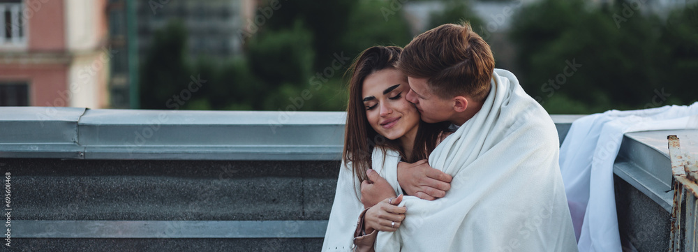 Beautiful young loving couple on a surprise romantic date on a roof top. Picturesque view, skyscrapers on background. Wine, candles, kisses on holiday Saint Valentine's Day celebration outdoors - obrazy, fototapety, plakaty 