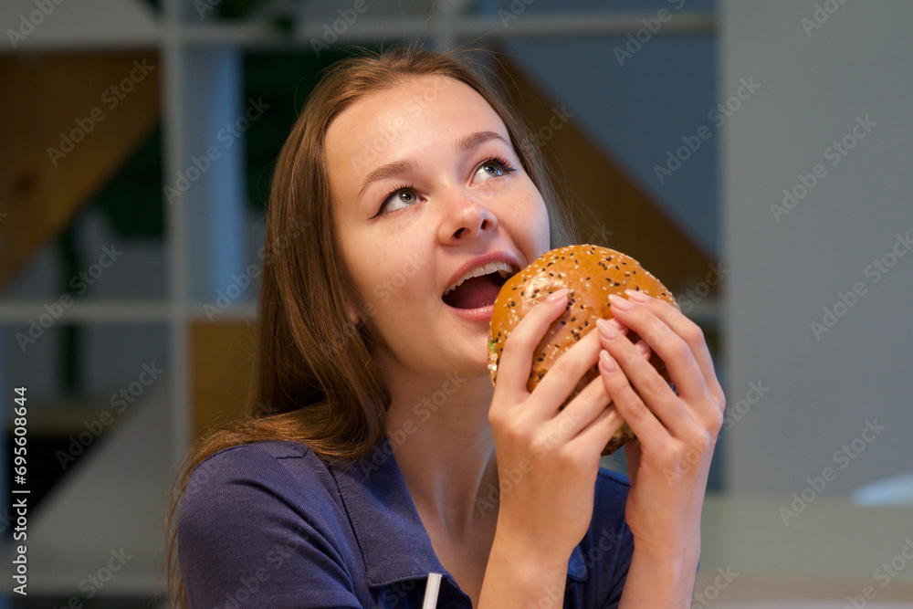 Portrait of young happy woman or beautiful teenager girl eating fast junk food, tasty burger and drink soda in a restaurant or cafe, enjoy meal. 