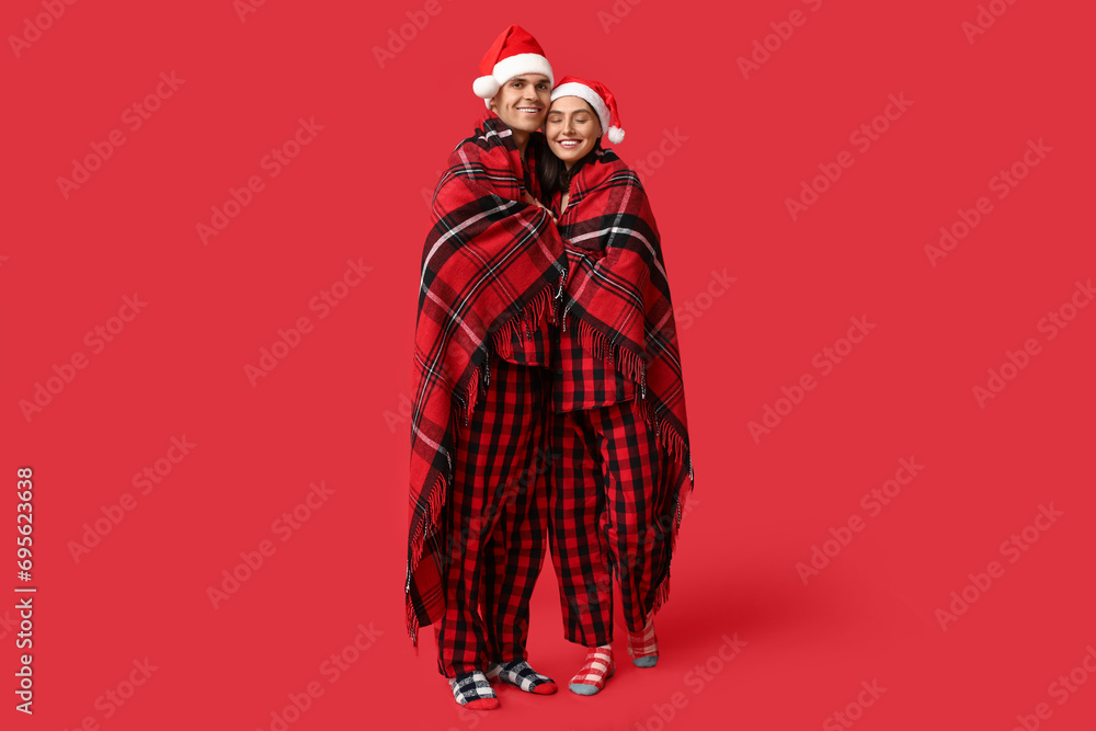Happy young couple in Christmas pajamas and Santa hats wrapped in warm plaid on red background