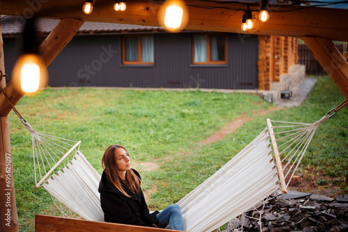 Beautiful young woman in black jacket relaxing on hammock on the backyard of wooden house cabin