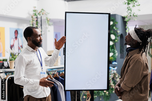 Clothing store assistant showing brand promotion on digital screen to customer. Mall worker advertising shoes new collection and offering client to check options on interactive white display photo