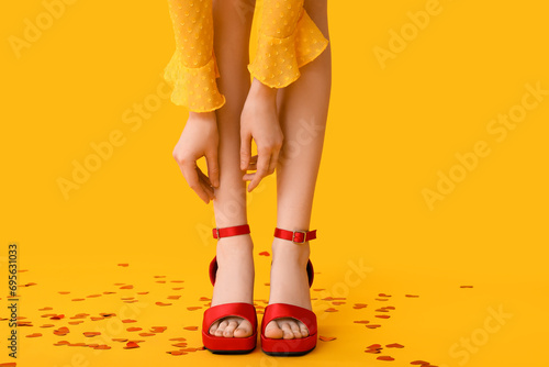 Legs of beautiful young woman in stylish high heels with confetti in hearts shape on yellow background