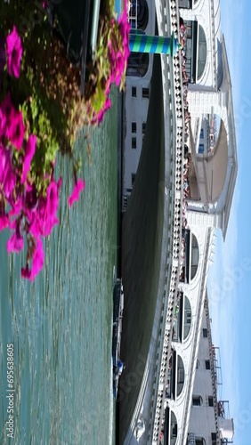 The rialto bridge with flowers planted in it on the foreground,a boat is passing under a bridge in sunny clear day