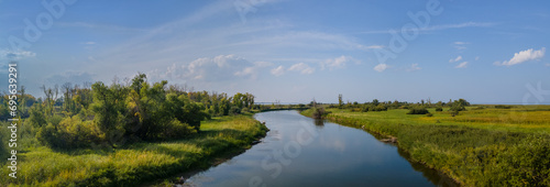 Panoramic view along a quiet curving river that is surrounded by green grass and small shrubs under a summer light-blue sky with scattered clouds. 