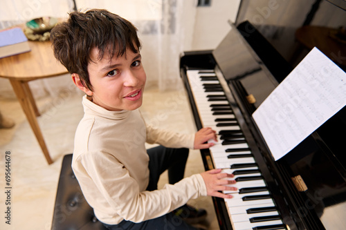 Confident authentic teenage boy smiles looking at camera, playing grand piano, composing music and tracks at home