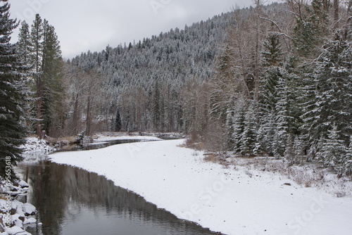 A slow flowing river in winter, with banks covered in snow, and snow covered trees and mountains in the background.