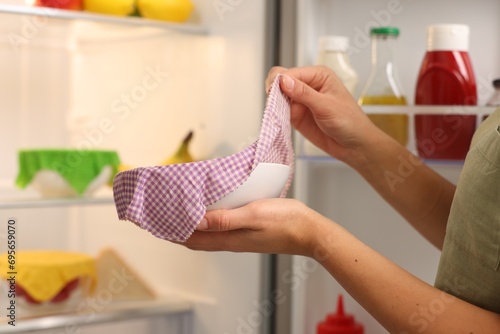 Woman taking away beeswax food wrap from bowl near refrigerator in kitchen, closeup