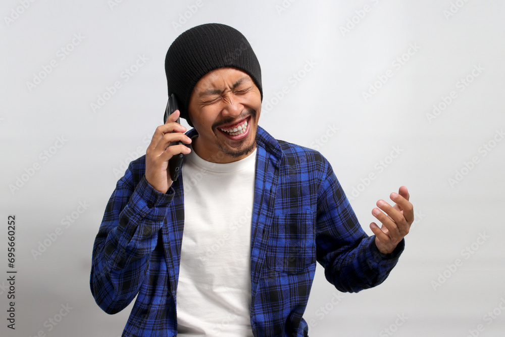 Cheerful young Asian man, dressed in a beanie hat and casual shirt, engages in a phone conversation, laughing and thoroughly enjoying a pleasant conversation while standing against a white background Cheerful young Asian man, dressed in a beanie hat and casual shirt, engages in a phone conversation, laughing and thoroughly enjoying a pleasant conversation while standing against a white background