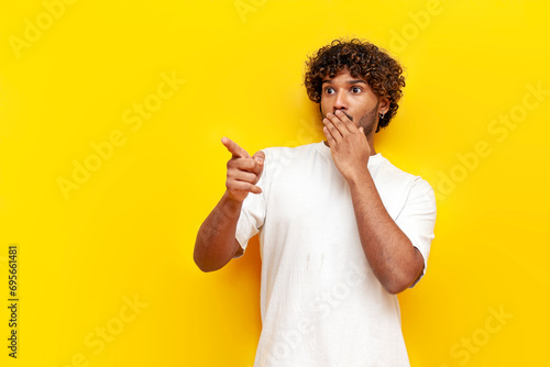 young indian shocked guy pointing in fear over yellow isolated background, surprised south asian man in white t-shirt photo