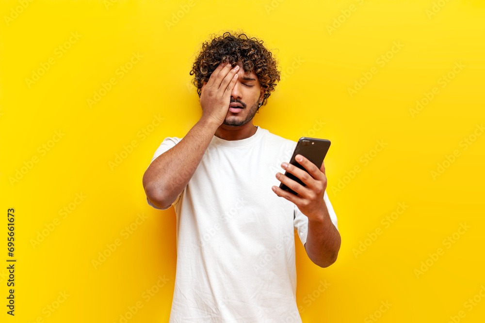 young indian sad guy using smartphone online with facepalm on yellow isolated background - obrazy, fototapety, plakaty 
