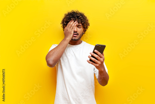 young indian sad guy using smartphone online with facepalm on yellow isolated background photo