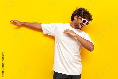 young indian guy in sunglasses dancing dab on yellow isolated background, south asian man in white t-shirt