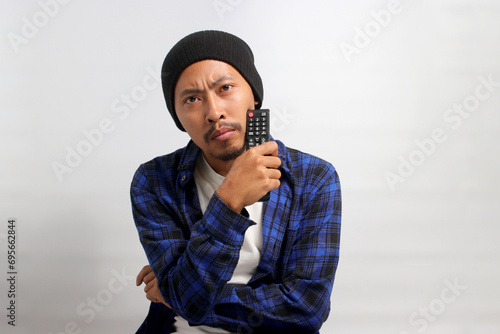 Pensive young Asian man, dressed in a beanie hat and casual shirt, appears puzzled while selecting a movie to watch, holding a TV remote at home, while standing against white background