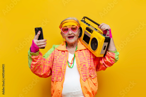 crazy old grandmother with tape recorder and smartphone in hipster clothes listens music on yellow background photo