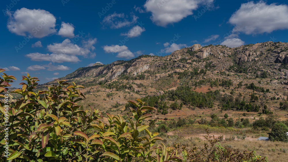 A mountain with rocky slopes against a background of blue sky and clouds. Green coniferous and deciduous trees grow on red soil. The landscape of Madagascar.