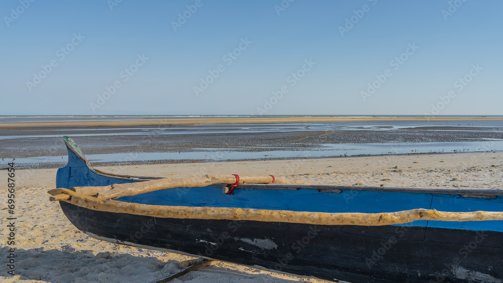 Traditional Malagasy pirogue on a sandy beach at low tide. The old wooden boat is painted in blue and black. Close-up. The ocean is visible far ahead. Clear blue sky. Copy space. Madagascar. Morondava