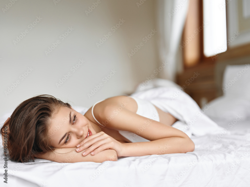 Young sexy woman in brunette lingerie lying in a white bed in the morning on a day off and smiling, vacation lifestyle, home waking atmosphere with sunlight from the window. Young sexy woman in brunette lingerie lying in a white bed in the morning on a day off and smiling, vacation lifestyle, home waking atmosphere with sunlight from the window.