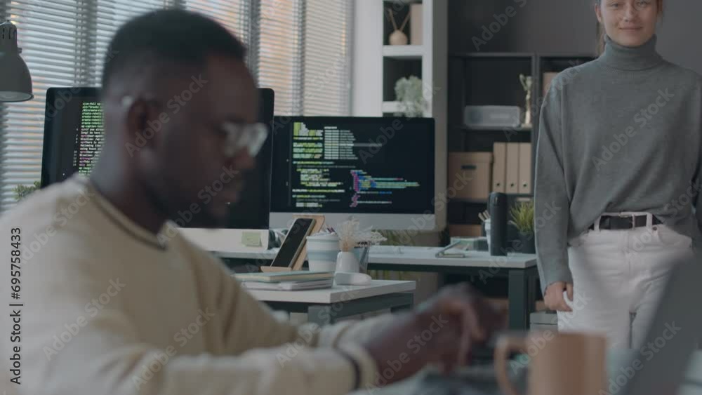 Medium shot of young Caucasian female programmer finishing coding on pc, getting up from her workplace and starting talking with Black male colleague, working together in modern office Medium shot of young Caucasian female programmer finishing coding on pc, getting up from her workplace and starting talking with Black male colleague, working together in modern office