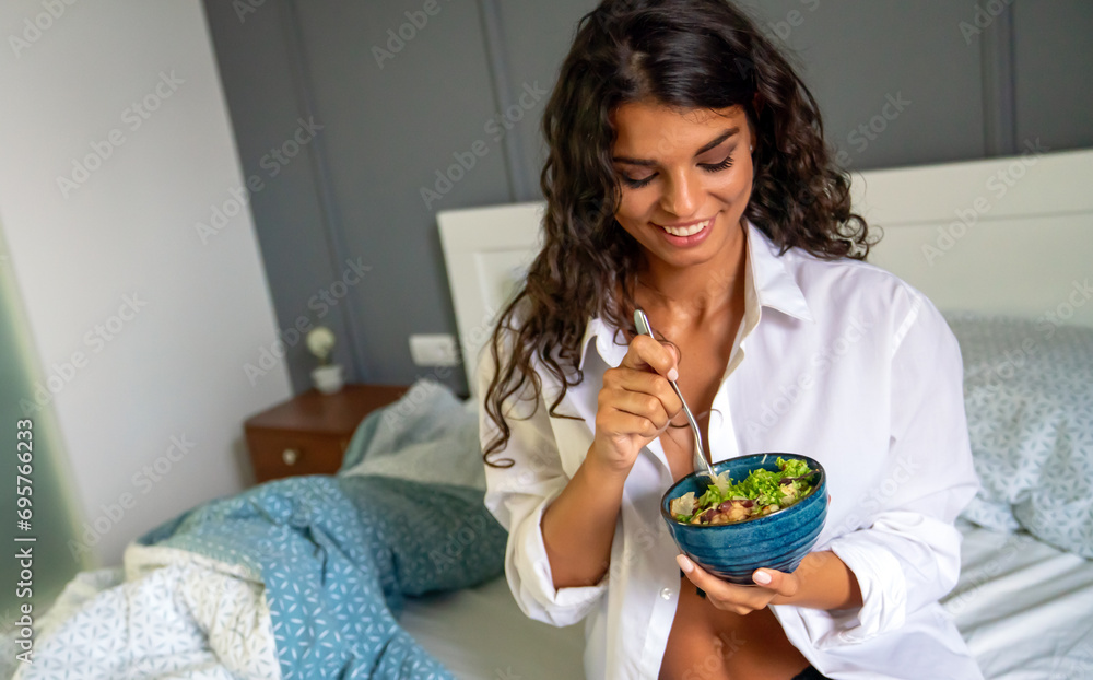 Portrait of a sexy young happy woman eating healthy salad in bedroom.