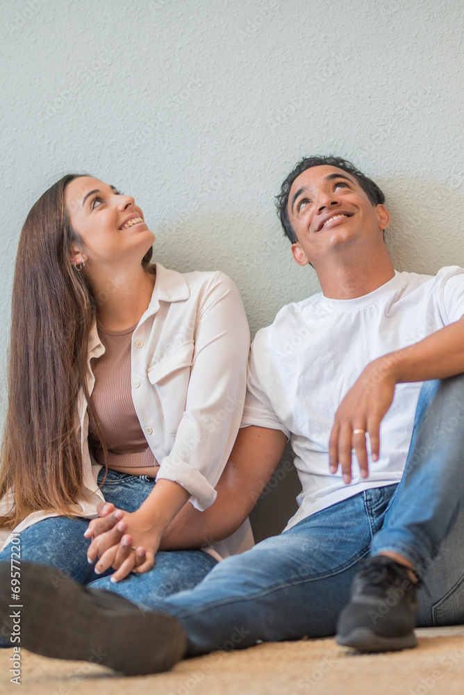Young couple at home sitting on the floor looking up in to the air and smiling - concept of love and relationship wito mortgage and new life together - interracial boy and girl Young couple at home sitting on the floor looking up in to the air and smiling - concept of love and relationship wito mortgage and new life together - interracial boy and girl