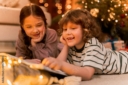 boy and girl have fun near Christmas tree  lay on floor reading a book near flashing lights  smiling and laughing during holidays