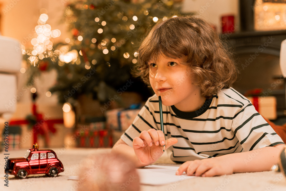 dark hair boy writes a letter with wish list to Santa Claus using color pencils laying on floor near Christmas tree.