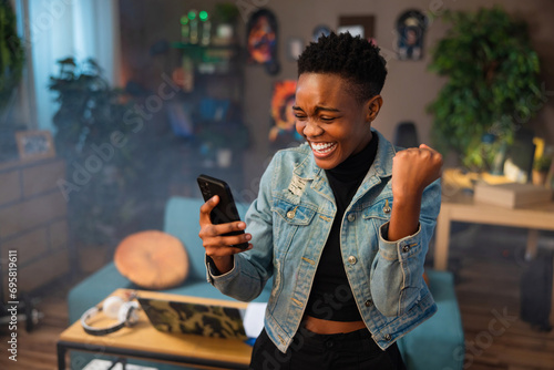 Happy african american teen girl lady woman with short hair using cell phone holding in hands smiling celebrating raising fists.