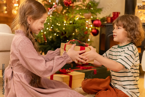 boy and girl sit near decorated Christmas tree, take presents and shake them to guess what is inside, laugh smile and are excited of foretaste photo