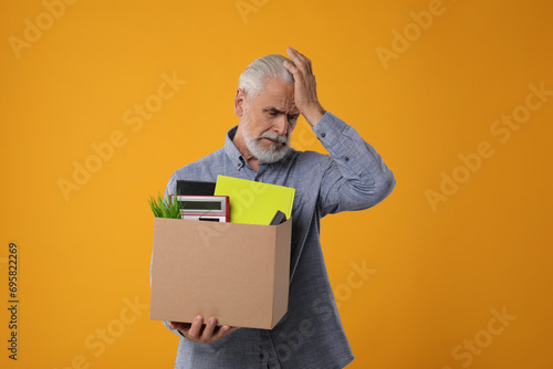 Unemployed senior man with box of personal office belongings on orange background