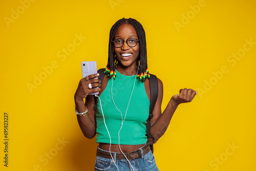 Young african feamle student, wears green top, braids, eyeglasses and backpack, listening to music on headphones and smartphone isolated over yellow background at studio. Technology concept. photo