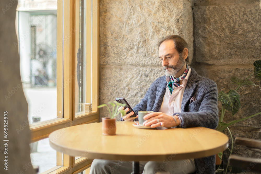 Senior man with beard using mobile phone sitting at a table in a cafe Senior man with beard using mobile phone sitting at a table in a cafe