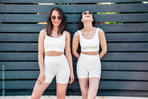Two young beautiful smiling female in summer white cycling shorts and top clothes. Sexy carefree women posing in street. Positive models having fun. Cheerful and happy. Near fence