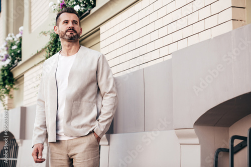 Handsome confident stylish hipster lambersexual model. Sexy modern man dressed in elegant white suit jacket. Fashion male posing in the street background in Europe city at sunset