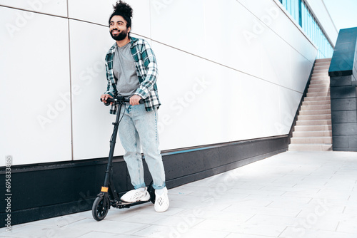 Trendy smiling bearded man in casual clothes riding electric scooter in urban background. Handsome model posing in the street near wall. Hipster guy with curly hairstyle. Cheerful and happy © halayalex