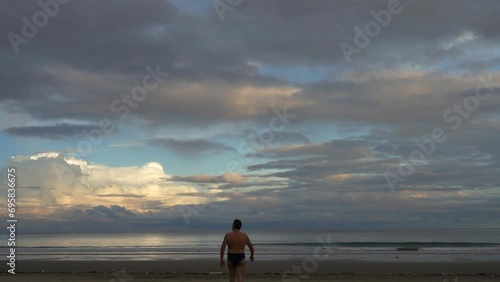 Early morning on a tropical island. The clouds in the blue sky are illuminated with golden light. A man walks towards the ocean on a sandy beach. The waves roll towards the shore, foaming. Malaysia. 