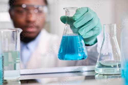 Close up shot of conical flask with blue liquid substance in hand of defocused African American laboratory specialist putting container on shelf