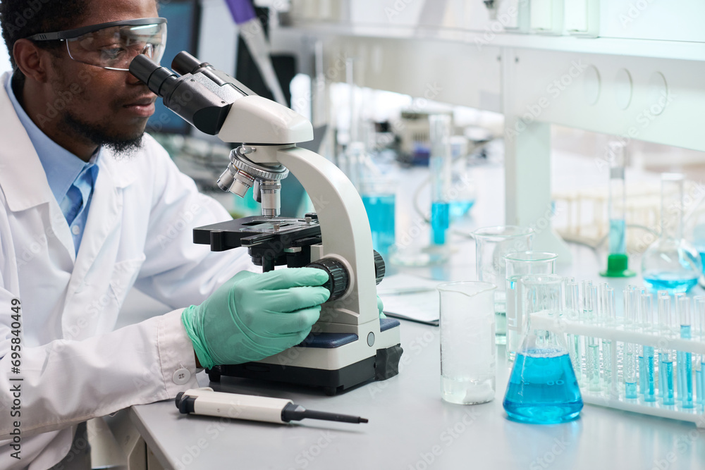 Side view at African American male scientist in safety goggles at laboratory desk with medical glassware studying samples with microscope Side view at African American male scientist in safety goggles at laboratory desk with medical glassware studying samples with microscope