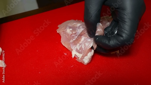 A gloved hand skillfully uses a knife on a red cutting board to trim a chicken fillet, removing the skin, hymen, and excess fat for a clean, ready-to-cook piece