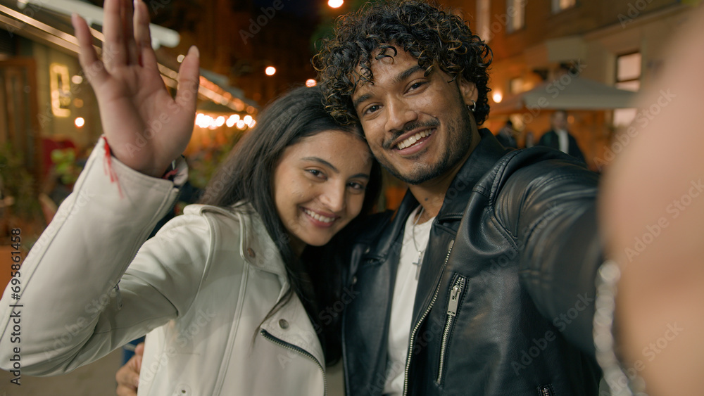 Selfie vlog travel blog influencer record video social media webcam view looking at camera waving hand greeting smiling happy Indian Arabian couple bloggers girl guy woman man in night evening city