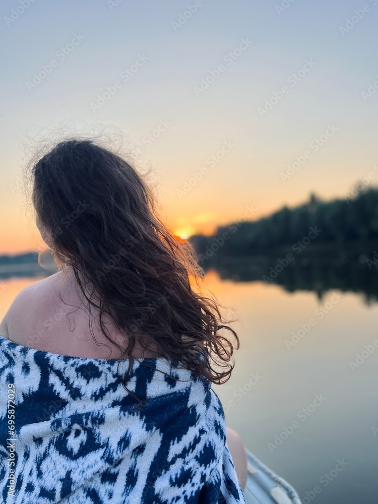 Photo of girl with loose hair from back on inflatable boat in evening on river among forest. Fishing. Vacation. Hobby. Twilight. Nature. Sunset. Sky. Water. Waves.