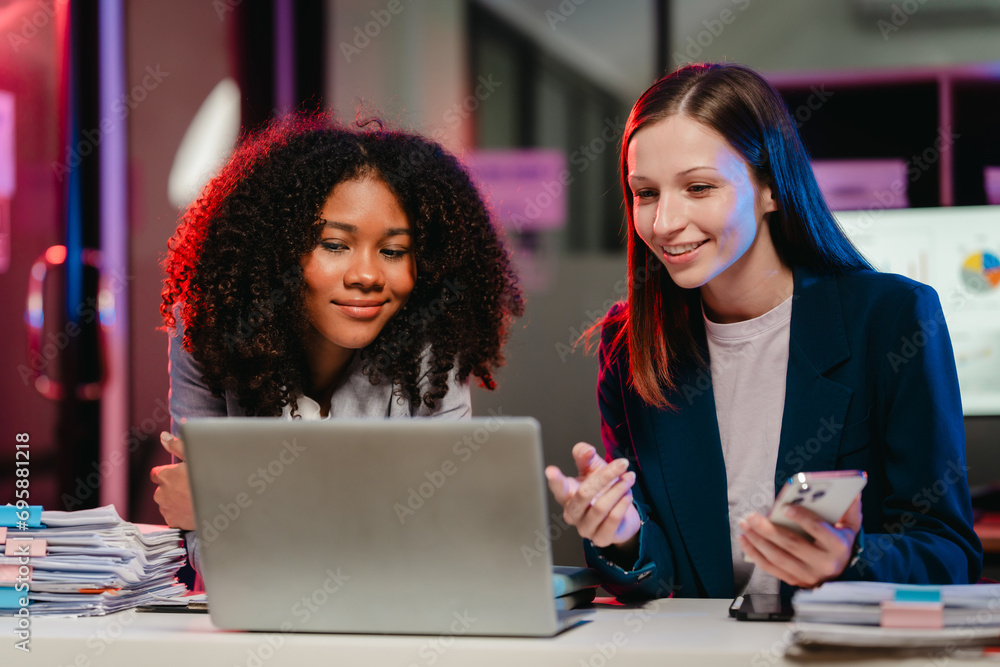 happy African American office worker with an afro and a Caucasian colleague collaborating over a laptop and documents at night. happy African American office worker with an afro and a Caucasian colleague collaborating over a laptop and documents at night.