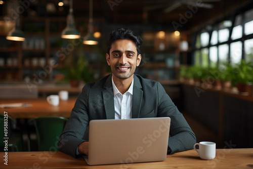 Smiling confident Indian business man employee looking at camera sitting at work desk with laptop computer. Portrait of smart happy businessman office worker or entrepreneur posing at modern workplace