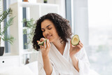 Close up portrait of young multinational female in robe holding two avocado halves in hands in bedroom interior. Young African American curly woman appreciating green avocado for great skin benefits.