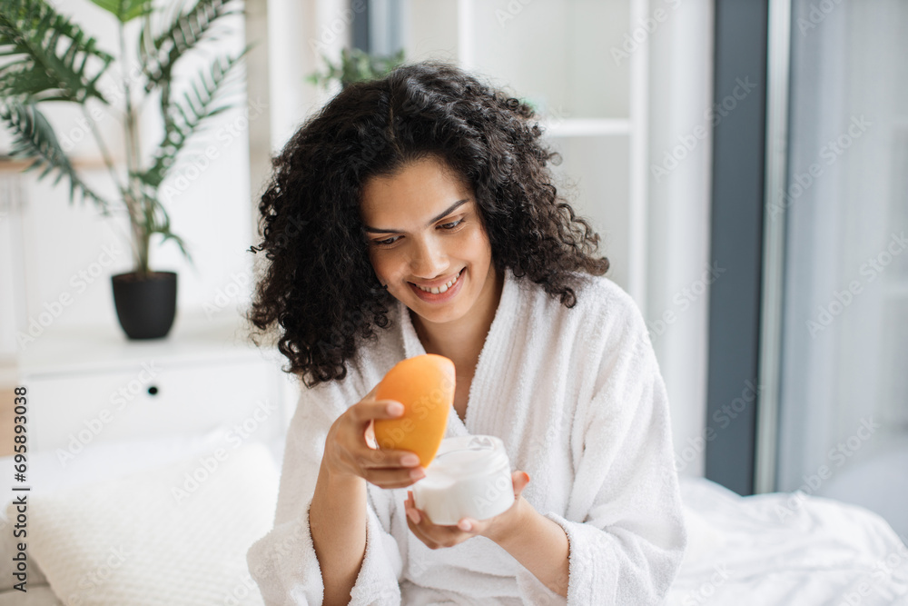 Happy young multinational woman in bathrobe holding half of grapefruit and making homemade cream for healthy skin on background of bright bedroom.