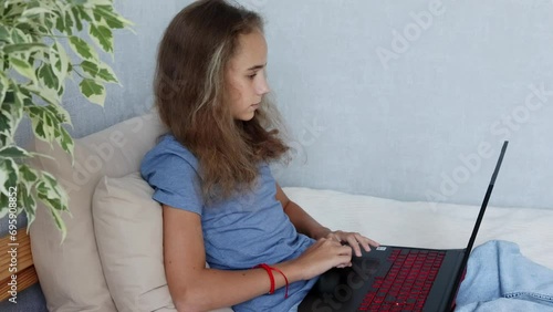 A blonde teenage girl is sitting on her bed typing on her laptop computer in her bedroom photo